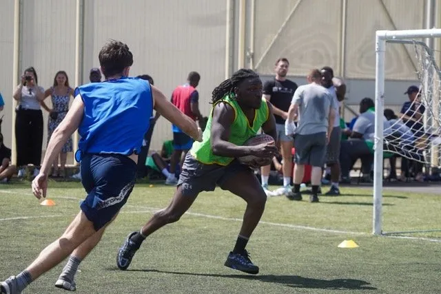 A dynamic rugby match on an outdoor field where a player in a green bib sprints forward with the ball while being pursued by a player in blue
