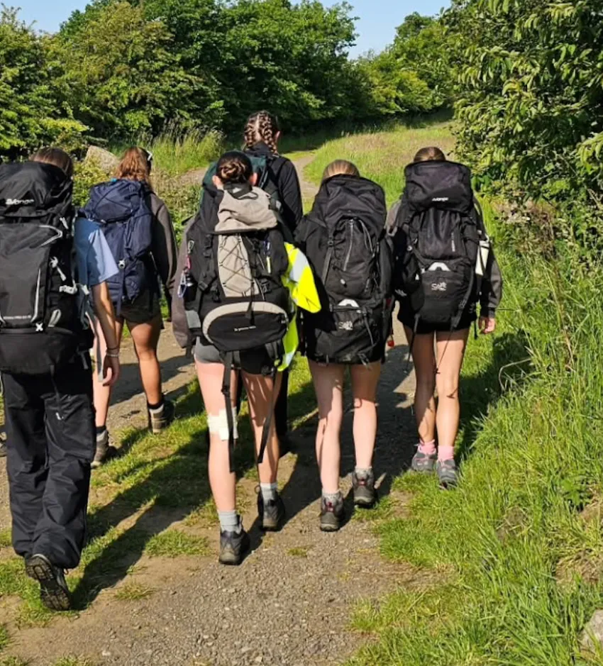 A group of young people wearing walking boots and carrying large rucksacks are walking away from the camera on a rural path