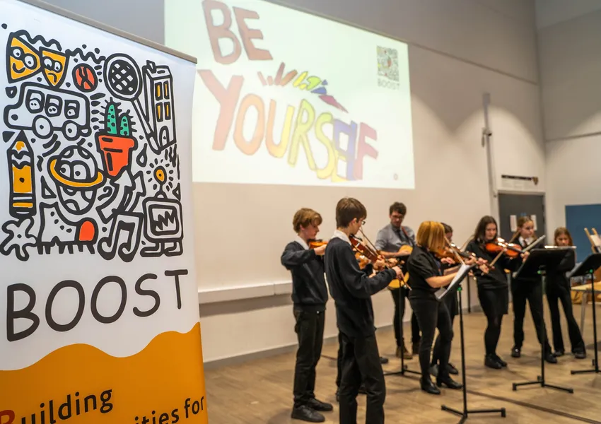 Students wearing school uniform are playing violins and other instruments. They are stood in front of a projection which says "Be Yourself" in colourful writing. In the foreground is a banner with the BOOST logo.