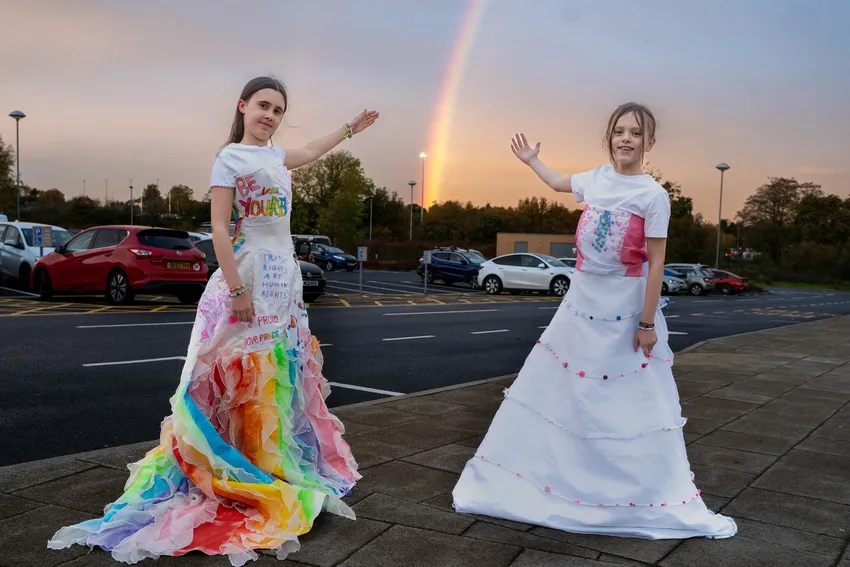 Two teenagers are wearing celebration dresses which are repurposed wedding dresses: one is decorated in rainbow colours and one is in the trans flag colours. The teenagers are standing in a carpark with a rainbow behind them.