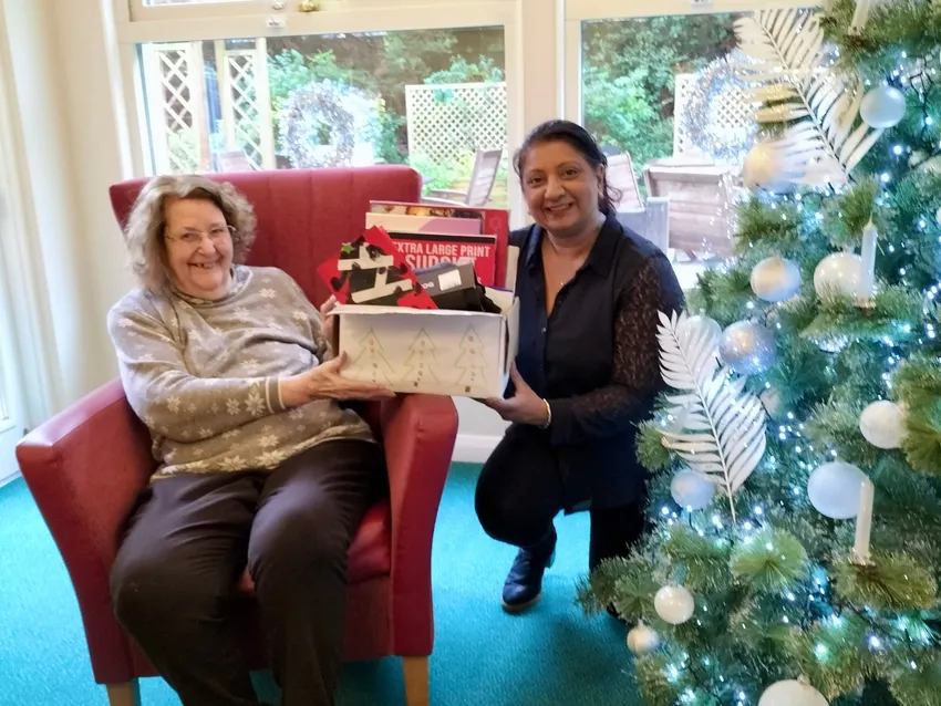 A care home resident sitting in a chair next to a Christmas tree. They are smiling and receiving a hamper of gifts from an Embracing Age representative.