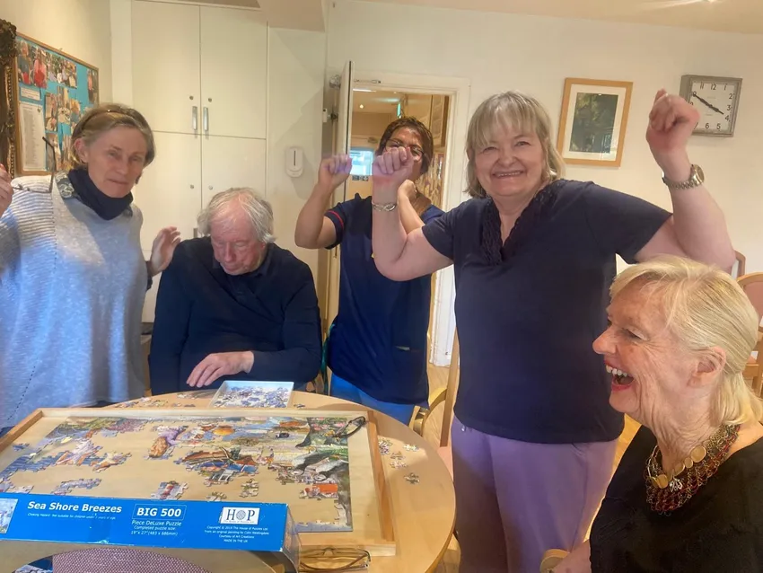 A group of care home residents and a volunteer are cheering and laughing while doing a jigsaw