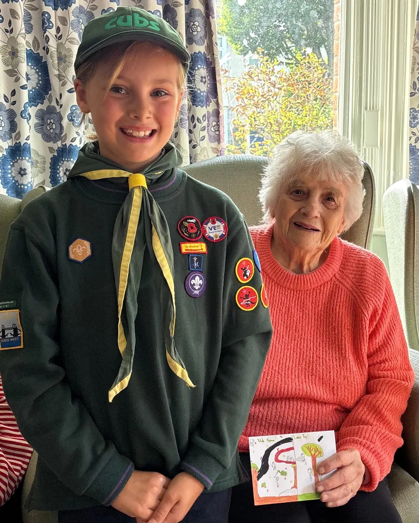 A child in Cub Scout uniform is standing next to a care home resident, who is holding a card drawn by the child. They are both smiling.