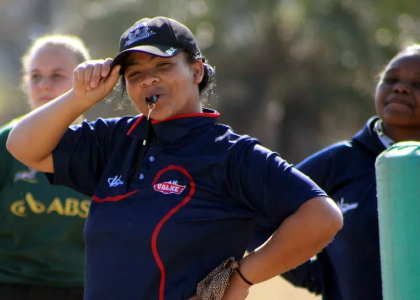 A rugby coach wearing a navy team polo shirt and cap blows a whistle while adjusting the brim of their hat during an outdoor training session.
