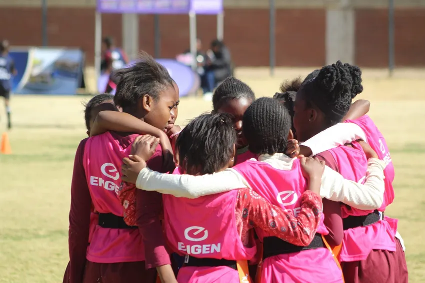 A group of young girls wearing bright pink sports bibs stand in a tight huddle with their arms around one another on a rugby field
