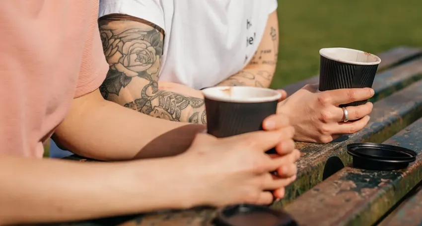 Two people sat next to each other, both holding a takeaway hot drinks mug in their hand.