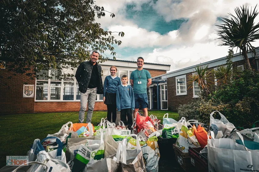 Two School Children, Their Teacher and a Member of Hospitality & Hope standing in front of a School. The sky is overcast and there are several bags of shopping containing donated goods assembled in front of them.