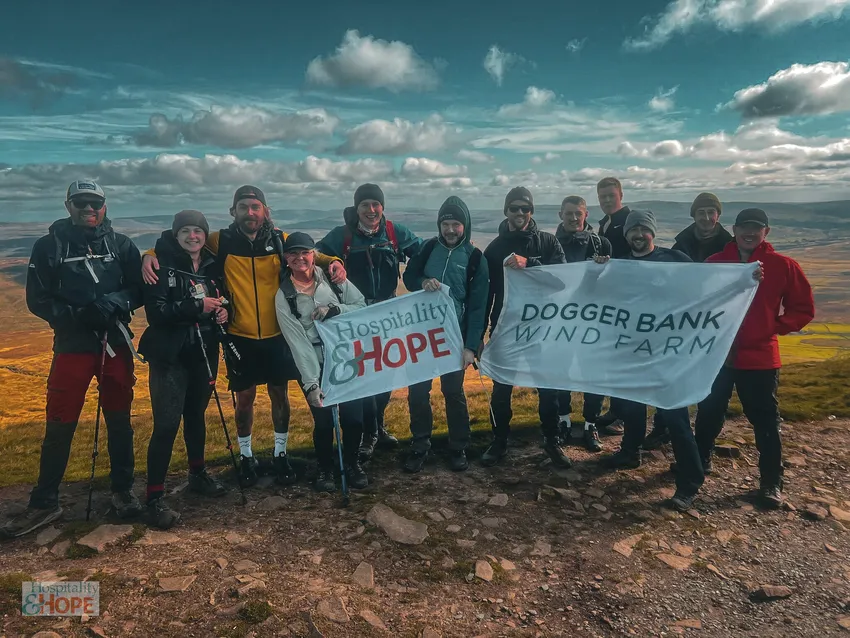 A group of hikers pose on a rocky hilltop with banners for Hospitality & Hope and Dogger Bank Wind Farm, with a wide landscape behind them.