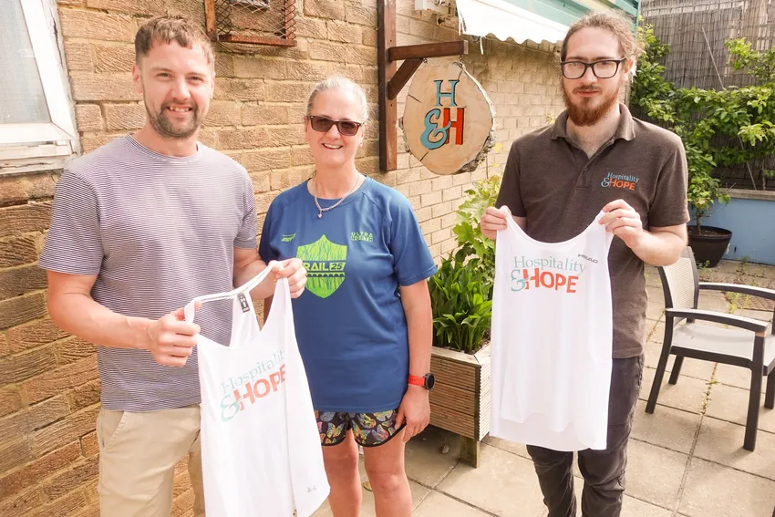Three people pose outside by a brick wall, with two holding white Hospitality & Hope vests.