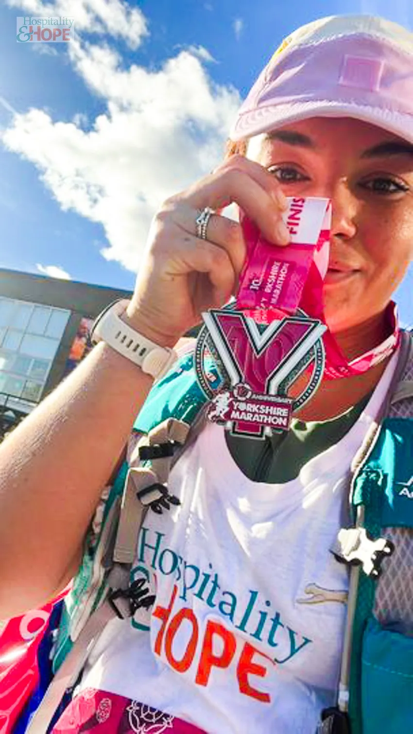 Close-up of a smiling runner wearing a Hospitality & Hope vest and holding up a Yorkshire Marathon medal outdoors.