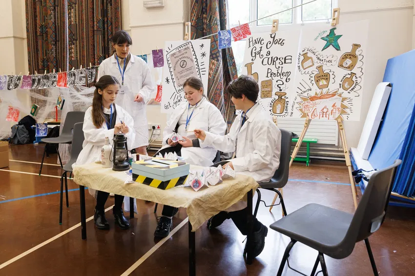 3 children wearing white lab coats, sat a table with creative materials. a female actor stands with them