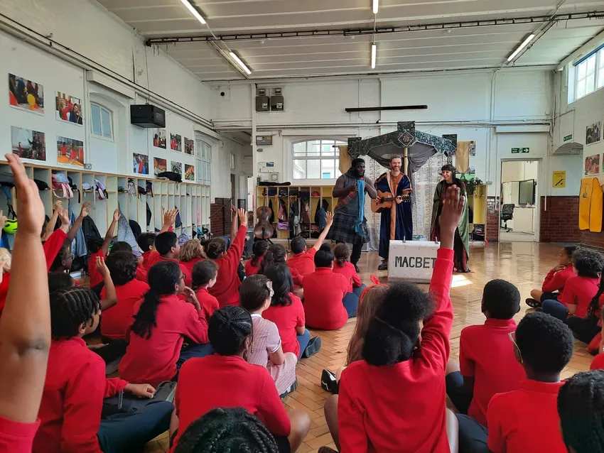 Three performers present a performance to a group of school children in a school hall, all with their hand up to ask questions