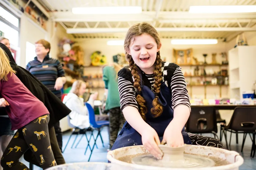 A smiling child throwing a pot on a potter's wheel.
