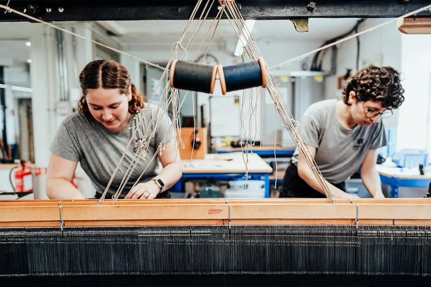 Weavers operate a historic loom at Whitchurch Silk Mill, visible through the narrow gap in the loom as they look over the harness of threads.