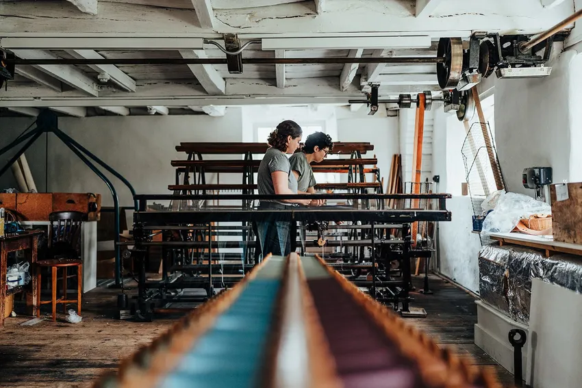 Two weavers stand beside a warping machine, inspecting threads. In the foreground, rows of silk bobbins are neatly arranged on a creel, with the large drum of the warping mill visible behind them.