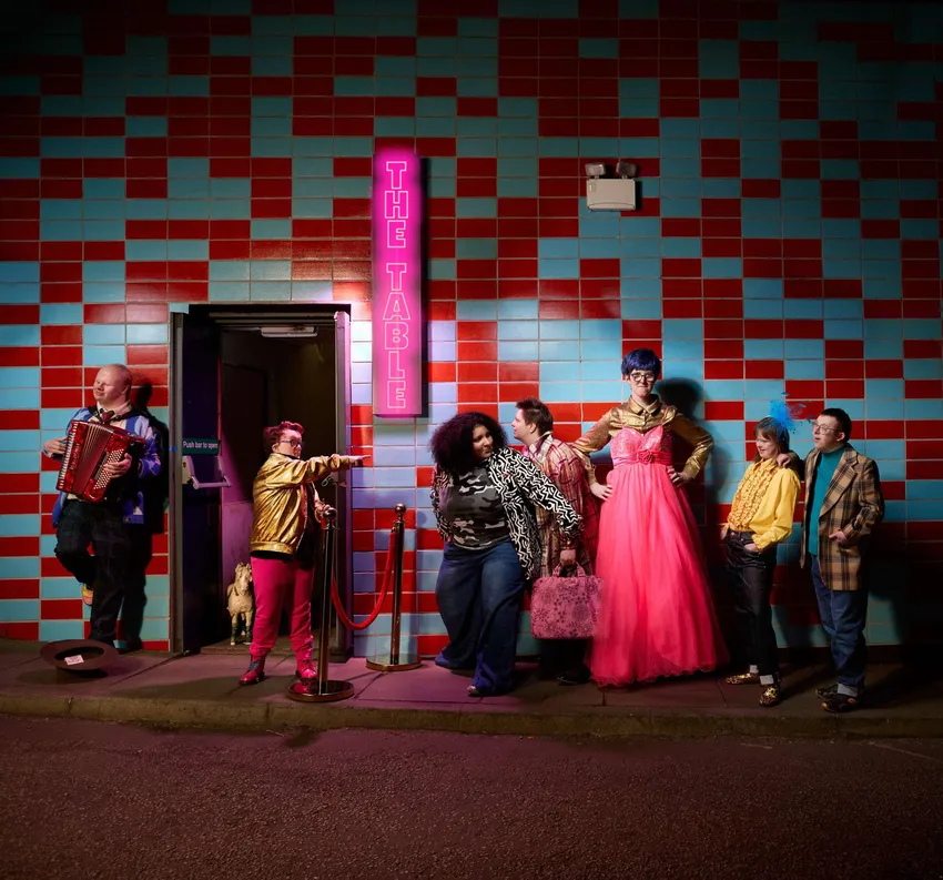 The Table, 2026. A group of people queue outside of a door. A wall in the background features red and blue tiles. A neon pink sign reads 'The Table'